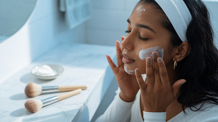 Indian woman smoothing sunscreen over her face before makeup, with makeup brushes on the counter