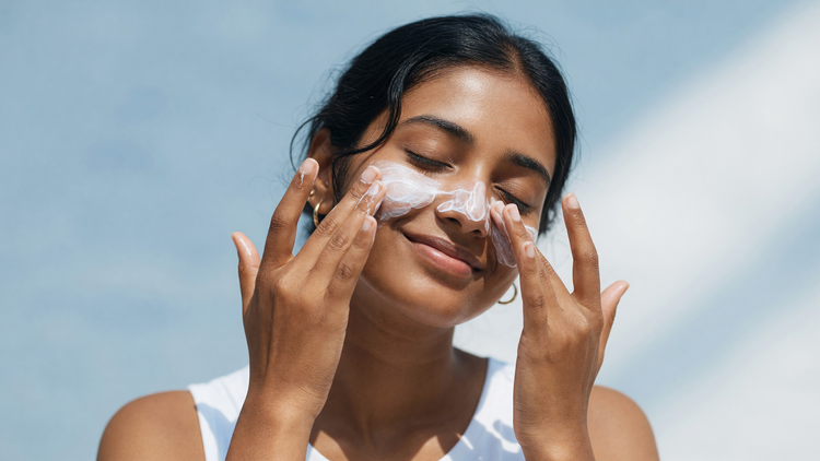 An Indian woman with warm brown skin smoothing sunscreen over her face outdoors under a bright blue sky
