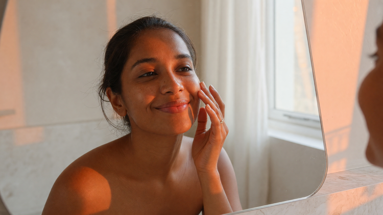 Indian woman with warm brown skin admiring her clear, even-toned complexion in a mirror in warm natural light